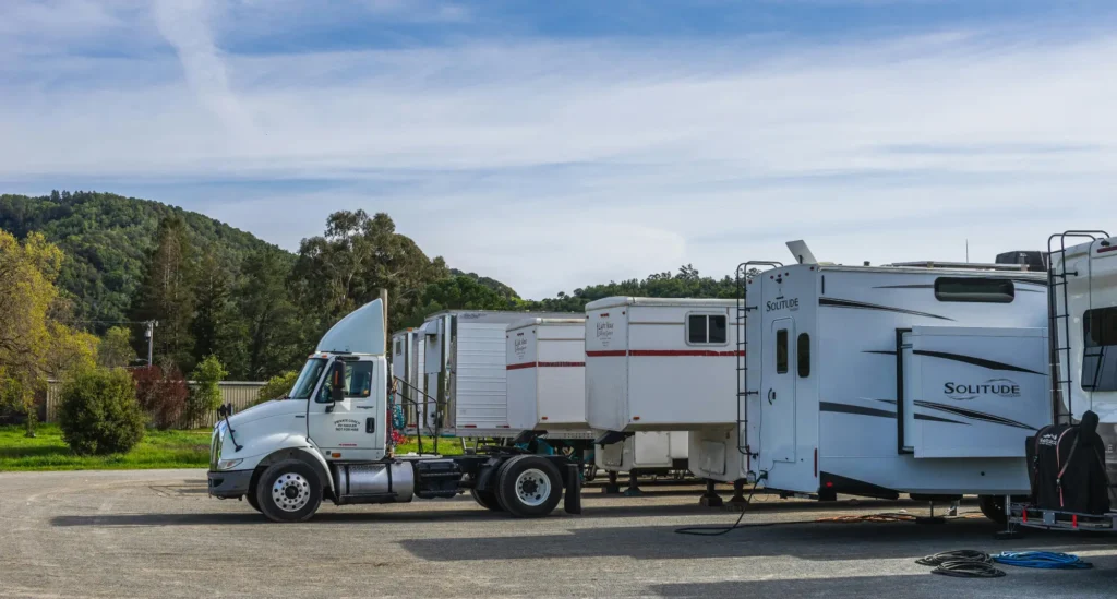 Semi-truck and travel trailers parked in an outdoor lot with green trees and mountains in the background under blue sky