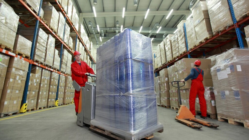 Two warehouse workers in red overalls moving wrapped pallets
