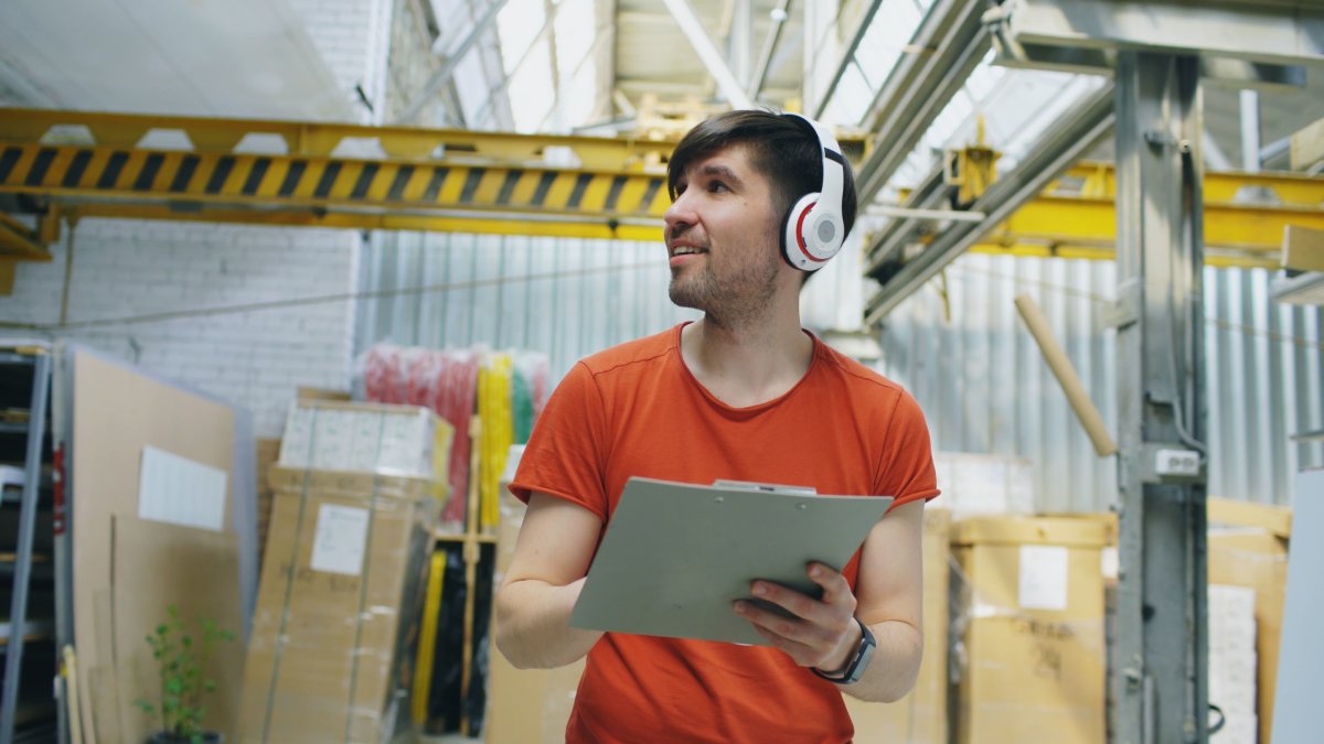 Young warehouse worker wearing headphones and holding a clipboard for inventory and tracking